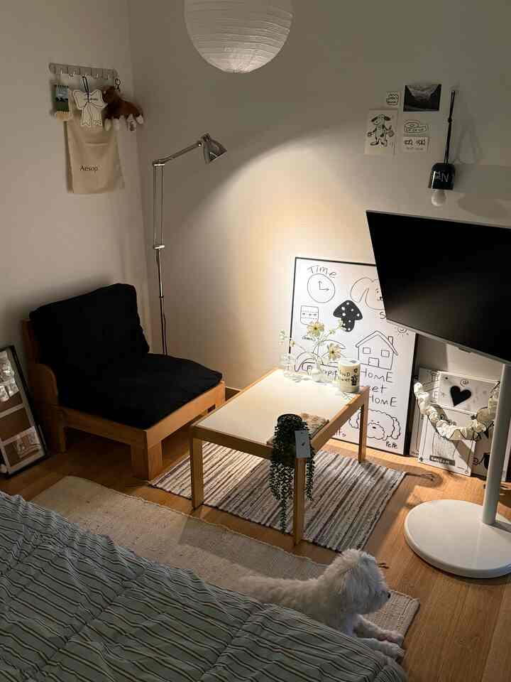ベージュとナチュラルウッド調の落ち着いた寝室、シンプルな黒クッションの椅子と多目的テーブルが配置され、白い犬が床に横たわる雰囲気のあるインテリア
