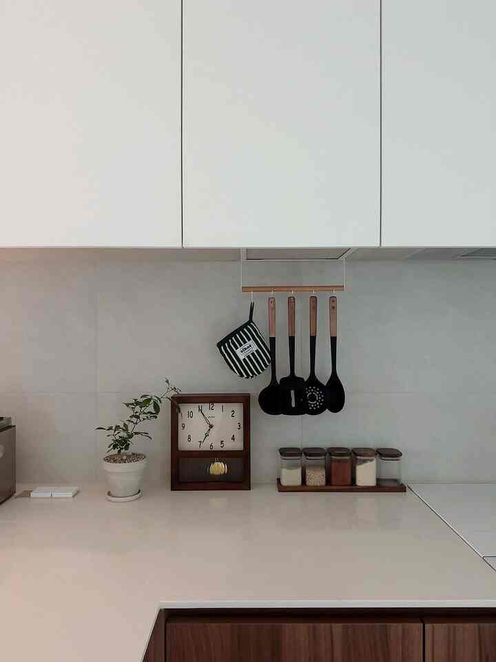 White-toned kitchen space featuring a wooden clock, hanging kitchen utensils, and a potted plant with a clean natural vibe