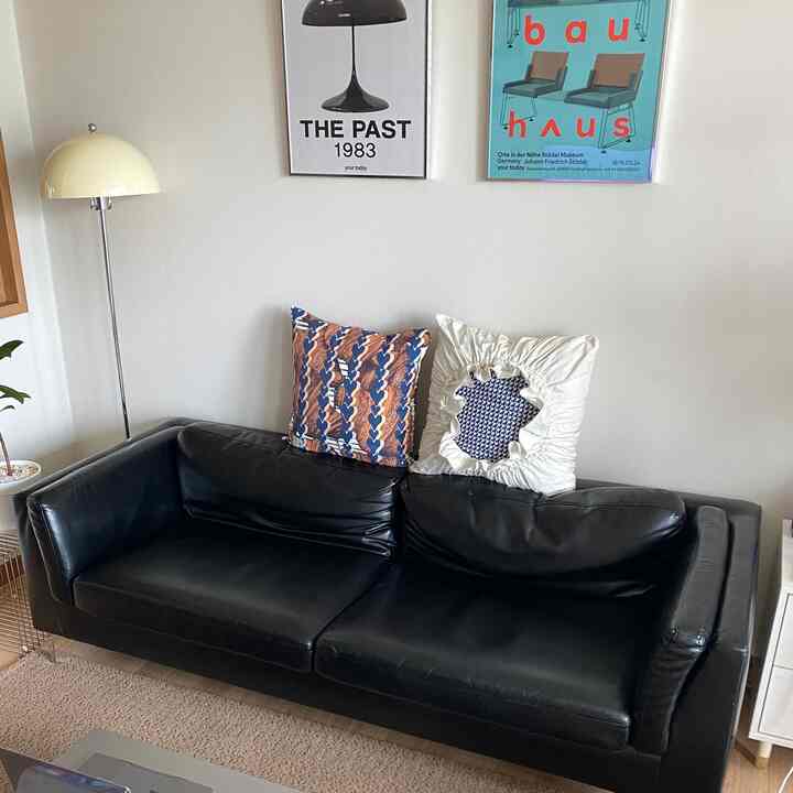 Simple living room with a black leather sofa featuring two cushions, modern framed posters on wall, and a yellow floor lamp