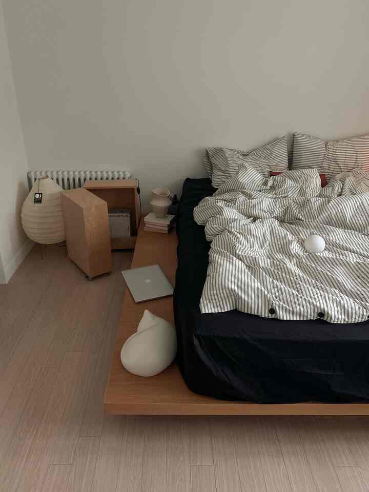 A bedroom with white walls and beige wooden floor featuring a black mattress with striped bedding on a natural wood bed frame, simple and natural atmosphere