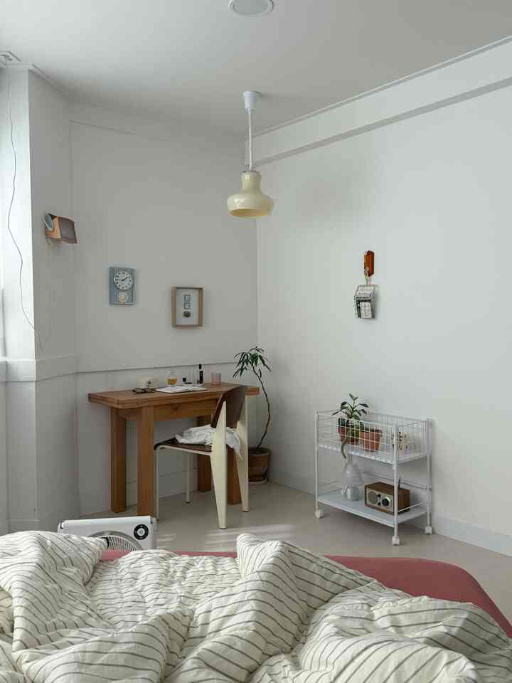 白と茶色の落ち着いた寝室、自然光が差し込む室内に木製のデスクと椅子、植物が配置されたシンプルで雰囲気のあるインテリア