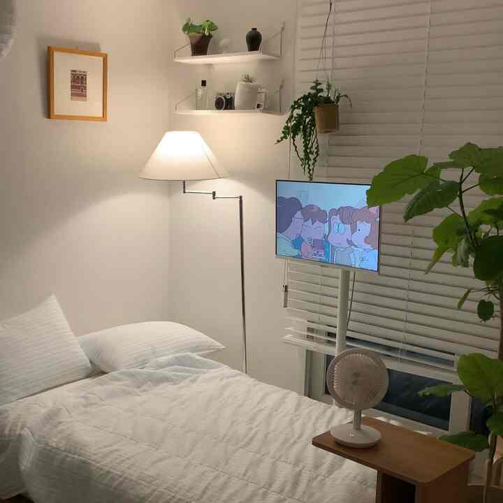 ホワイト調と木目調の調和したワンルーム寝室、整ったベッドとフロアランプ、移動式テレビスタンドが落ち着いた雰囲気を演出