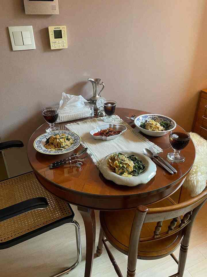 ベージュ色の壁とブラウンの丸いダイニングテーブルがある狭いリビング空間、パスタの皿と飲み物が配置されたナチュラルな雰囲気のインテリア