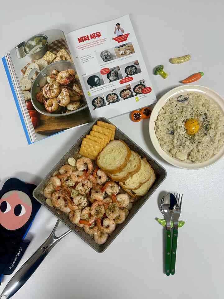 白いテーブルにチーズリゾットとバターシュリンプが入ったスクエアパン、レシピ本と調理器具が配置された明るく清潔なキッチンの雰囲気あるインテリア