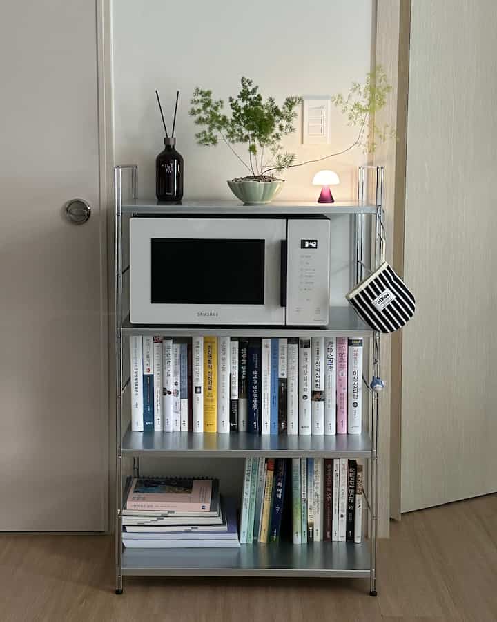 明るい木目調の床と白い壁のキッチン空間、スチール棚に電子レンジと本が整然と配置された雰囲気のあるインテリア