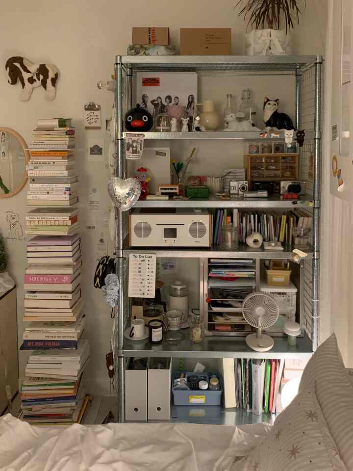 白とグレーのトーンの狭い寝室、中央に金属製の棚と積まれた本が配置された落ち着いた空間