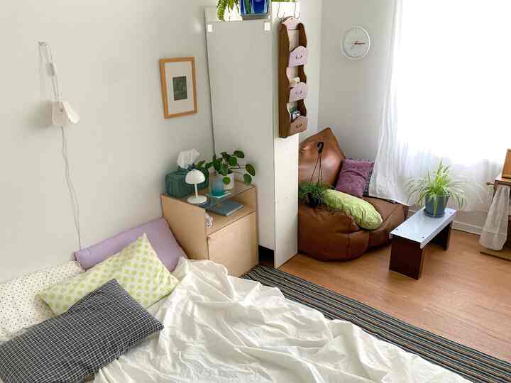 明るいナチュラルな色調の横長の4坪寝室、白いベッドとビーズクッションソファ、植物とカーテンが配置されたシンプルな空間