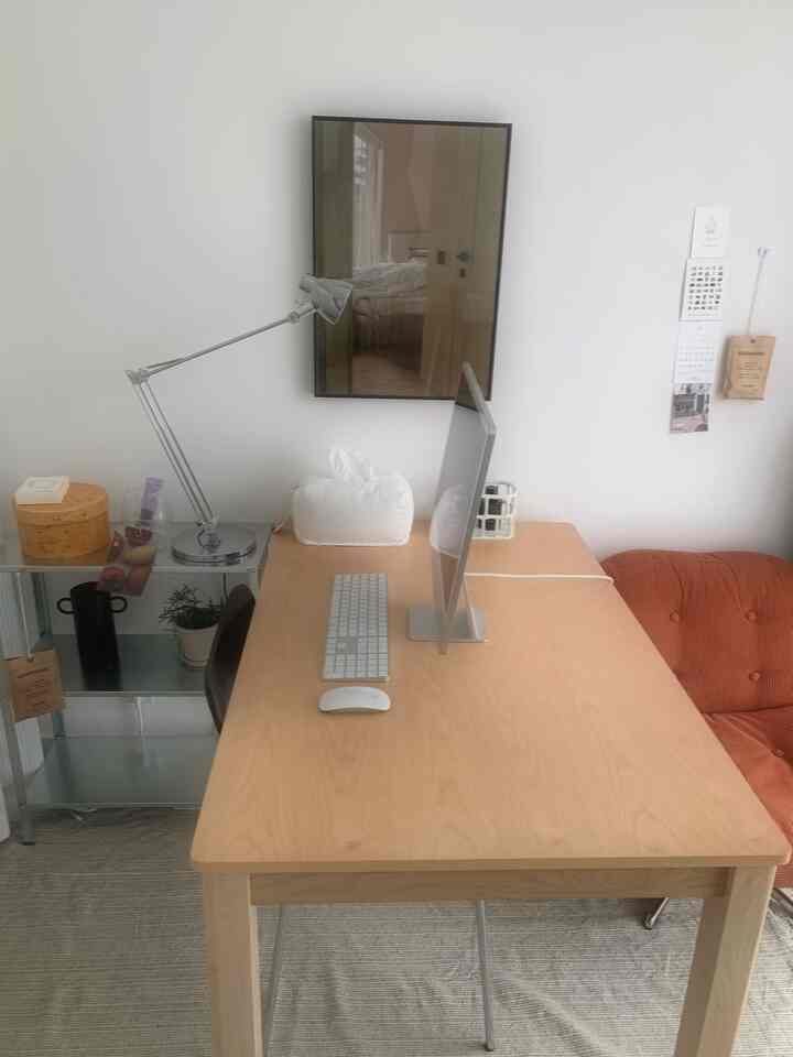 ライトブラウンの木製デスクが中心に配置されたモダンでナチュラルなリビングの作業空間、壁には額縁とイケアの棚がある