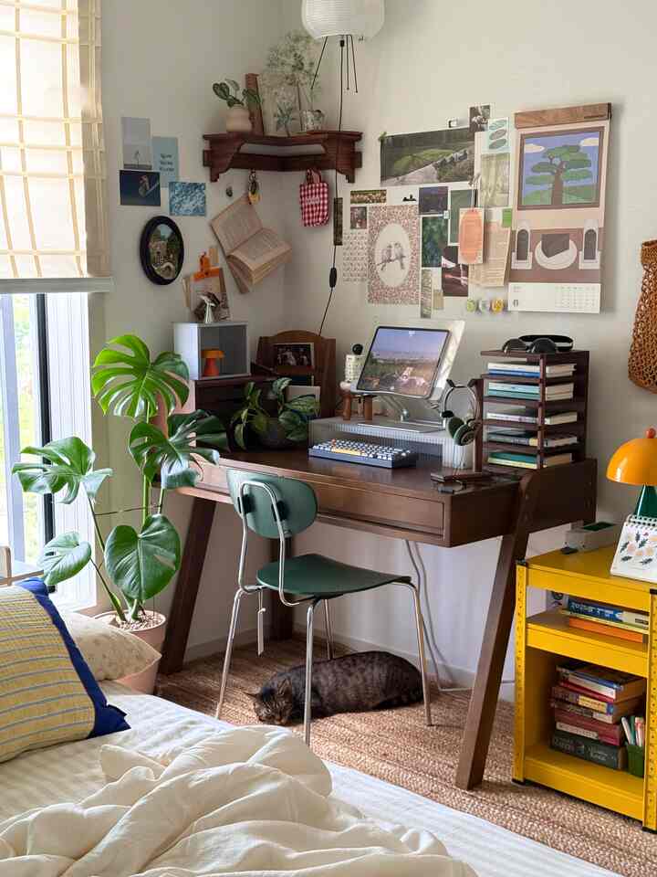 ナチュラルな木目調と緑の植物が調和した寝室兼ホームオフィス、ラグの上でくつろぐ猫がいる温かみのある空間