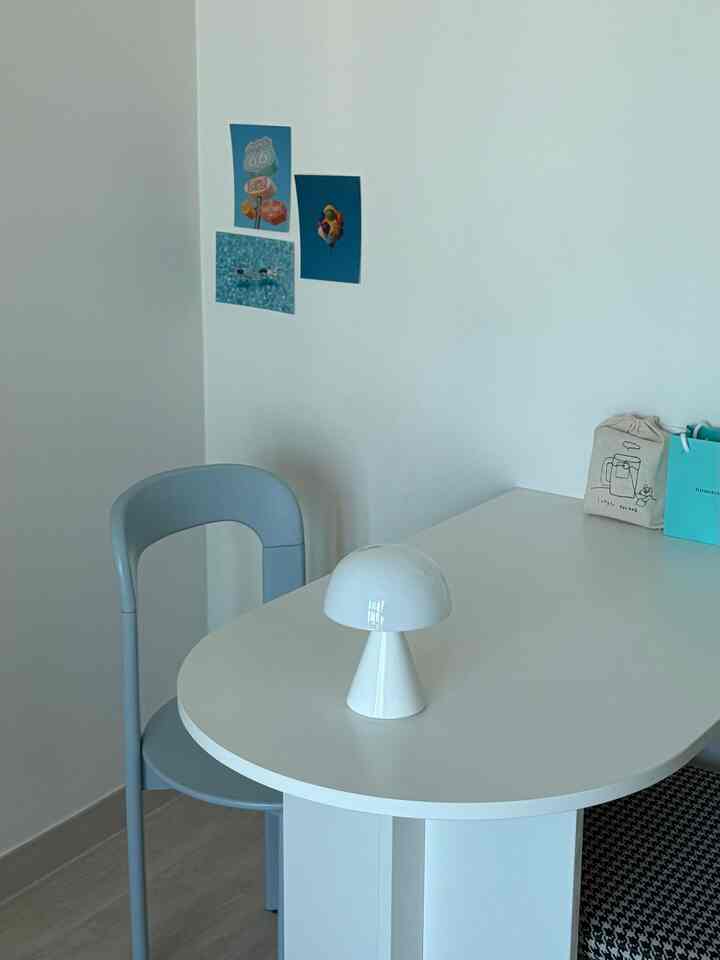 White and gray toned studio apartment featuring a simple dining table and chair with a table lamp and wall decor creating a modern space