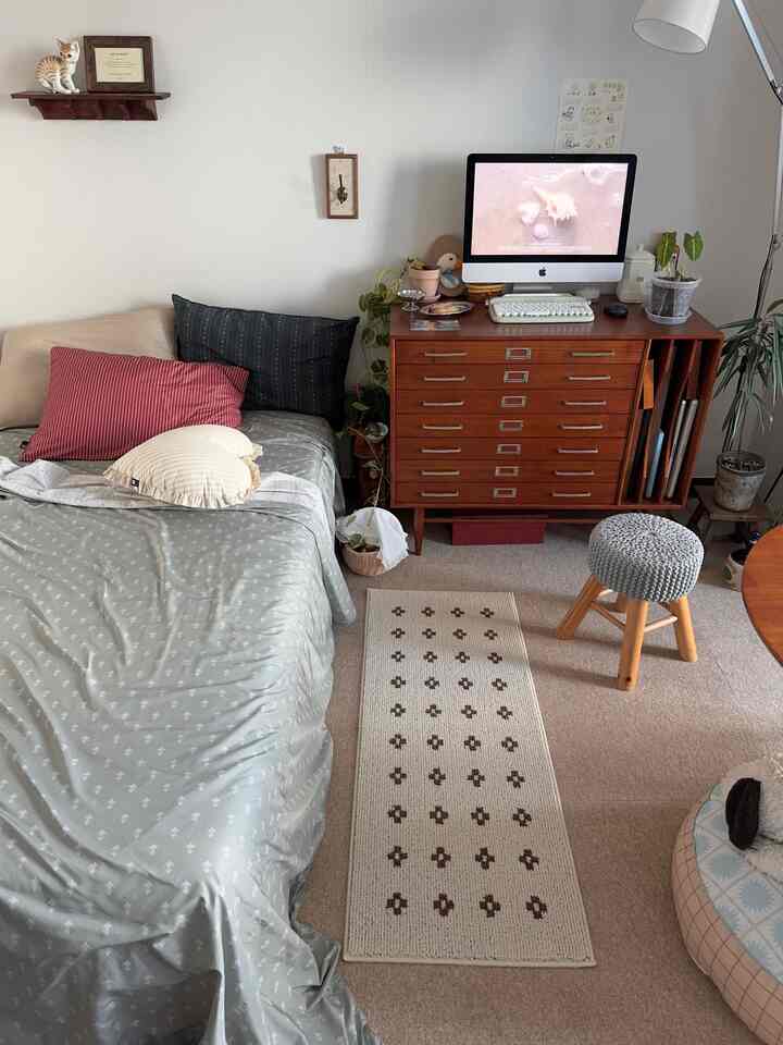 ナチュラルな色調の寝室、ウッドキャビネットにコンピューター、快適なベッドとラグが配置された雰囲気のあるインテリア