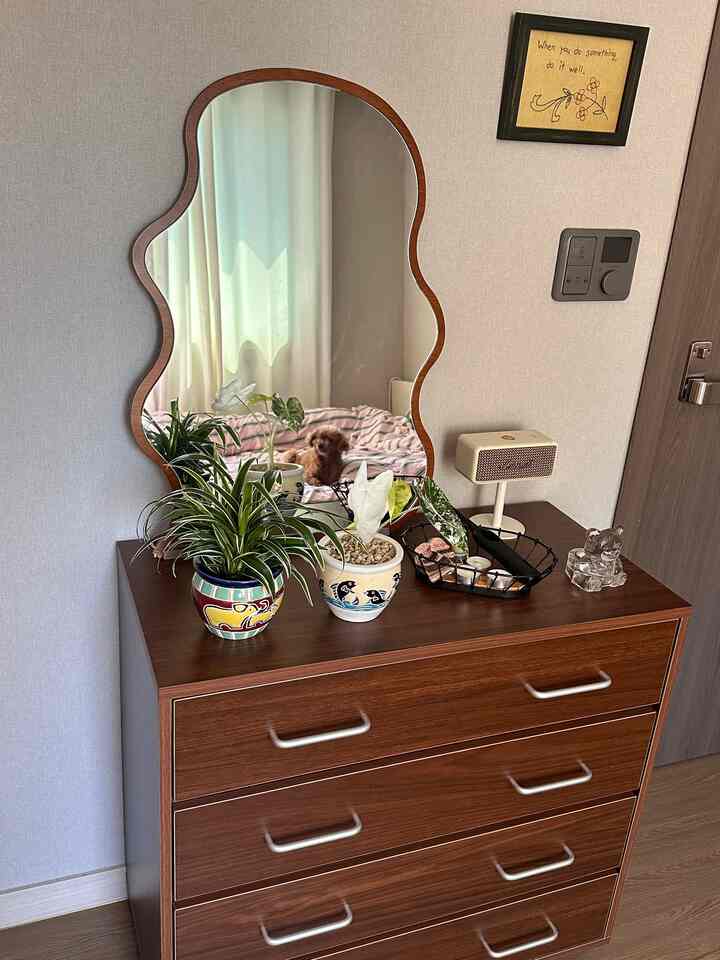 温かみのある木目調のチェストが置かれた寝室、曲線の鏡と植物で演出された雰囲気あるインテリア
