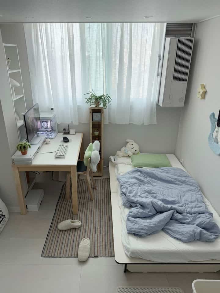 White-toned studio apartment featuring a desk with computer on the left and a bed on the right, creating a cozy and clean interior