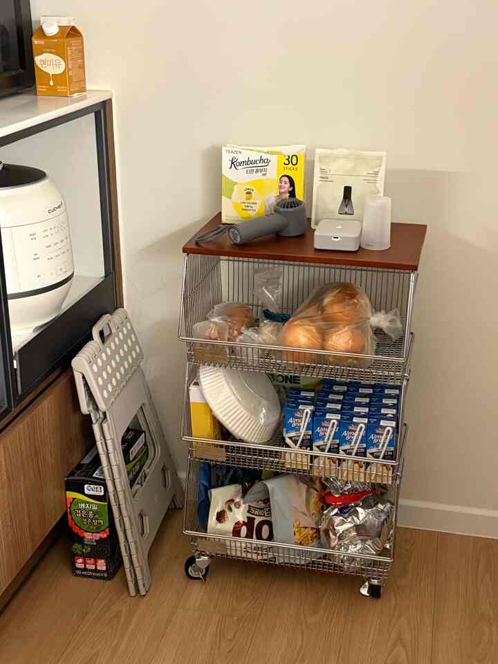 白い壁と木目調の床のあるキッチンの一角、皿や食材が置かれたメタルラックと折りたたみスツールが配置されたコンパクトな空間