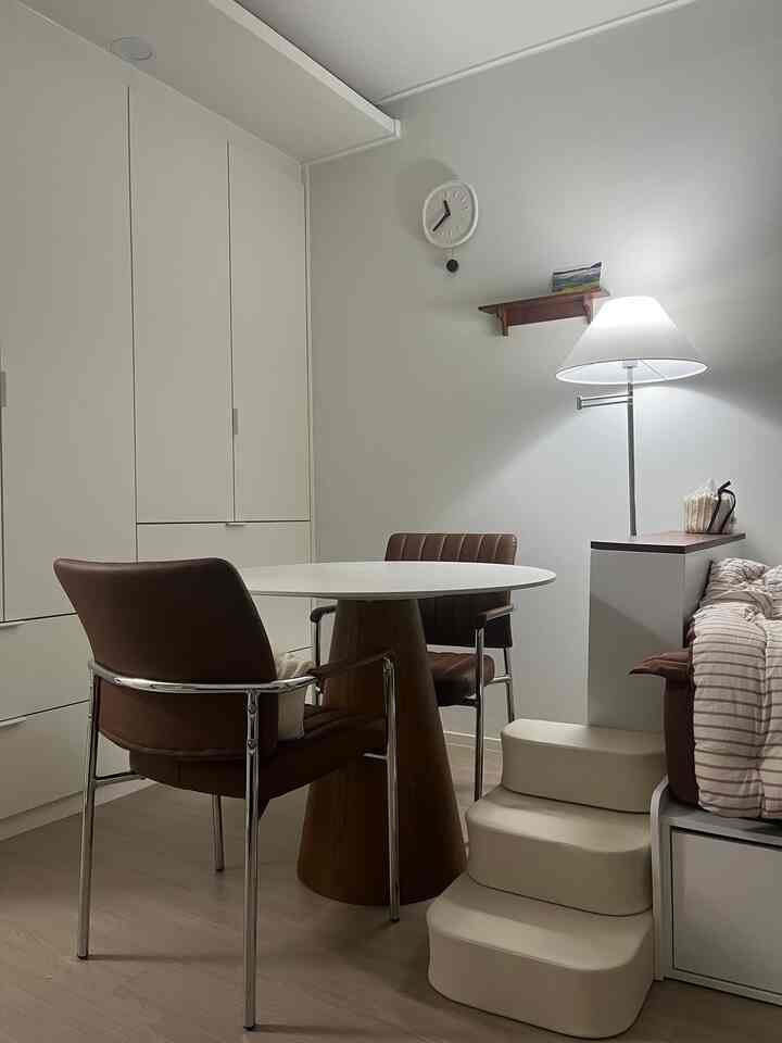 白と茶色のトーンのワンルーム、丸いダイニングテーブルとアームチェア、フロアランプが配置された雰囲気のある小さな部屋