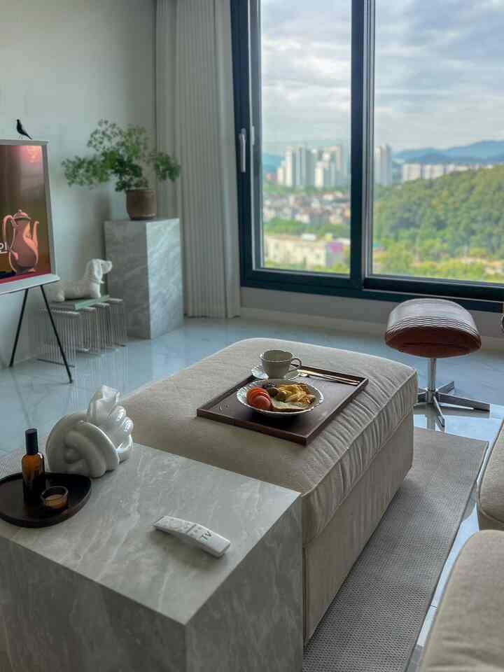 ナチュラルカラーのベージュ色のリビング、ベージュのソファと大理石のサイドテーブル、窓の外に街並みと緑が見える雰囲気のある空間