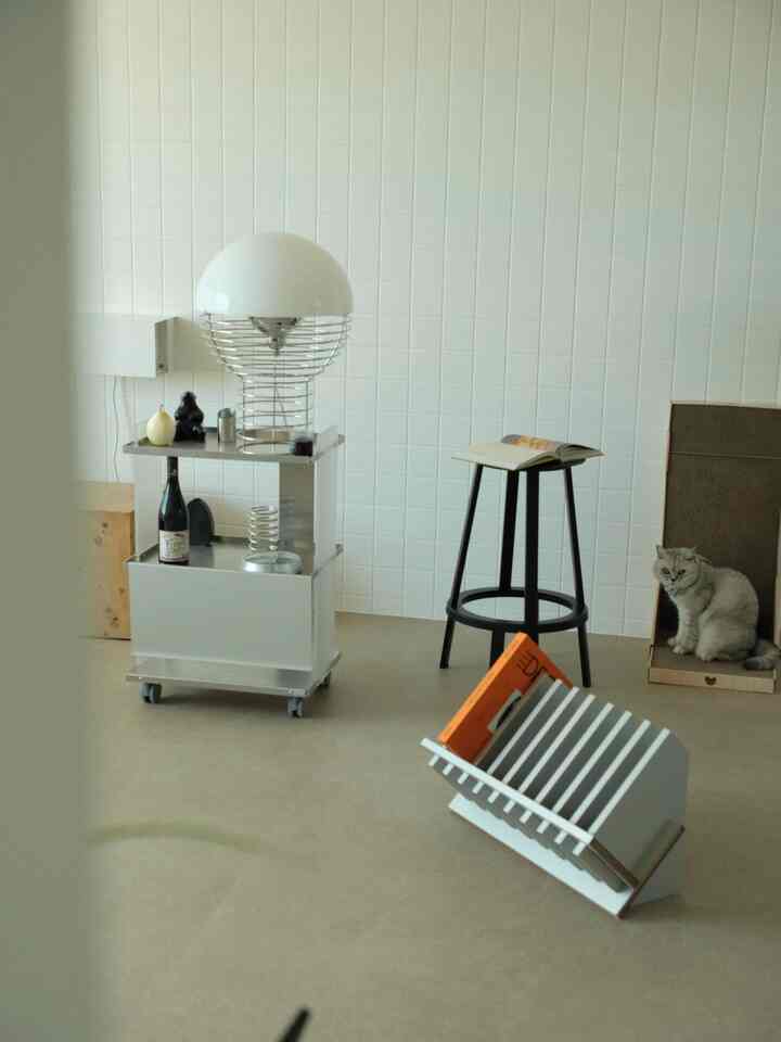 ホワイトタイルの壁とブラウンの床を持つ空間に、ホームバー用のスツールとバーカート、照明、そして猫と猫トイレが配置された印象的なインテリア