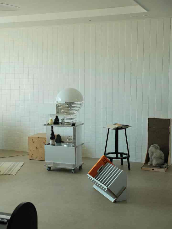 ホワイトタイルの壁とベージュの床があるミニマルなリビング、サイドテーブルとスツール、スクラッチャーのそばに猫がいるインテリア