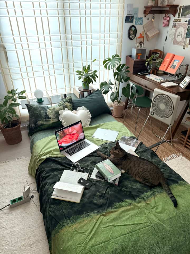 緑のトーンの寝室、植物と机が配置され、猫がベッドでくつろぐ雰囲気のあるインテリア