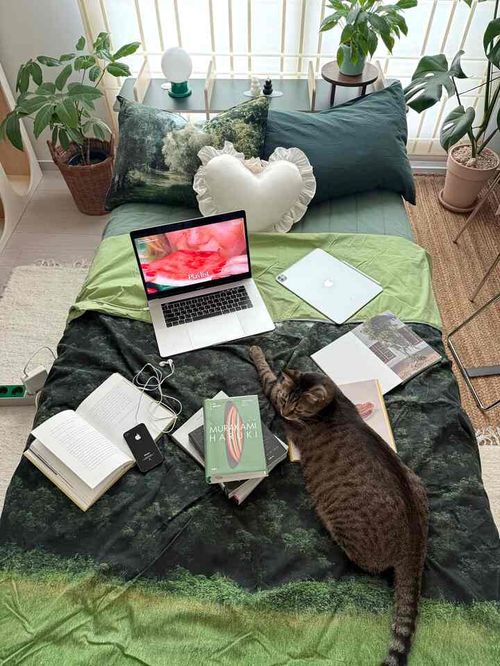 グリーントーンの寝室、植物と枕が置かれたベッドには猫や本、ノートパソコンが配され、落ち着いた雰囲気のインテリア