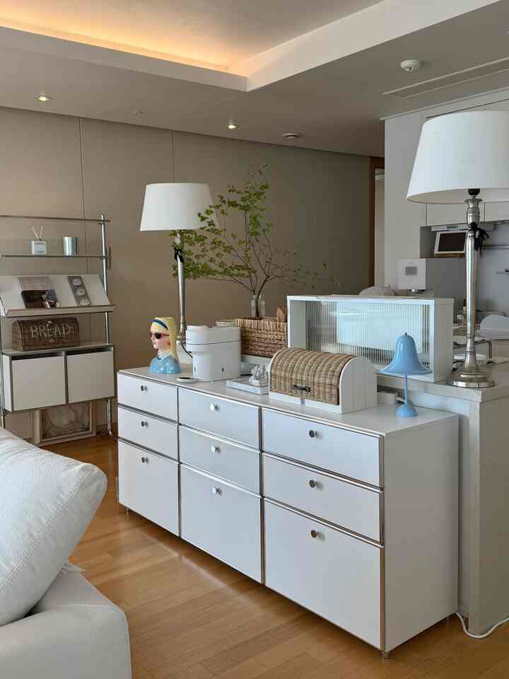 A living room with white and wood tones, featuring a large cabinet, floor lamps, and rattan accessories creating a neat and natural atmosphere
