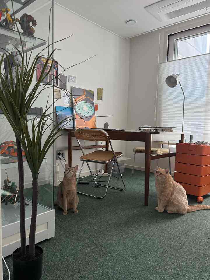 白い壁と緑のカーペットのロフト風書斎、デスクや椅子、フロアランプ、猫が二匹いる雰囲気のあるインテリア