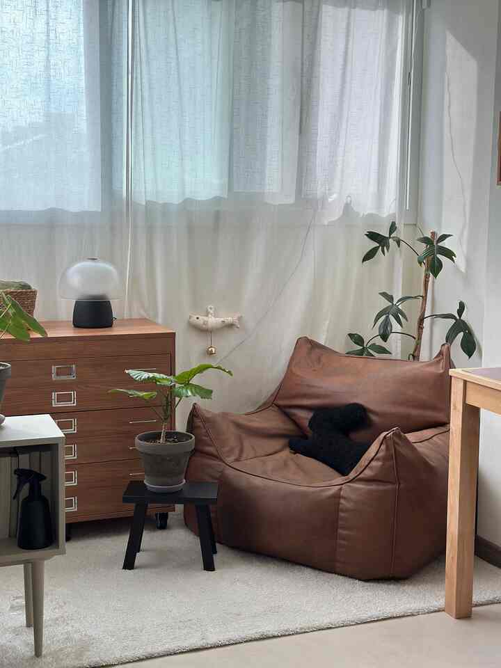 白いカーテンと木目調のチェスト、ブラウンのビーズクッションと植物が配置されたナチュラルモダンな寝室空間