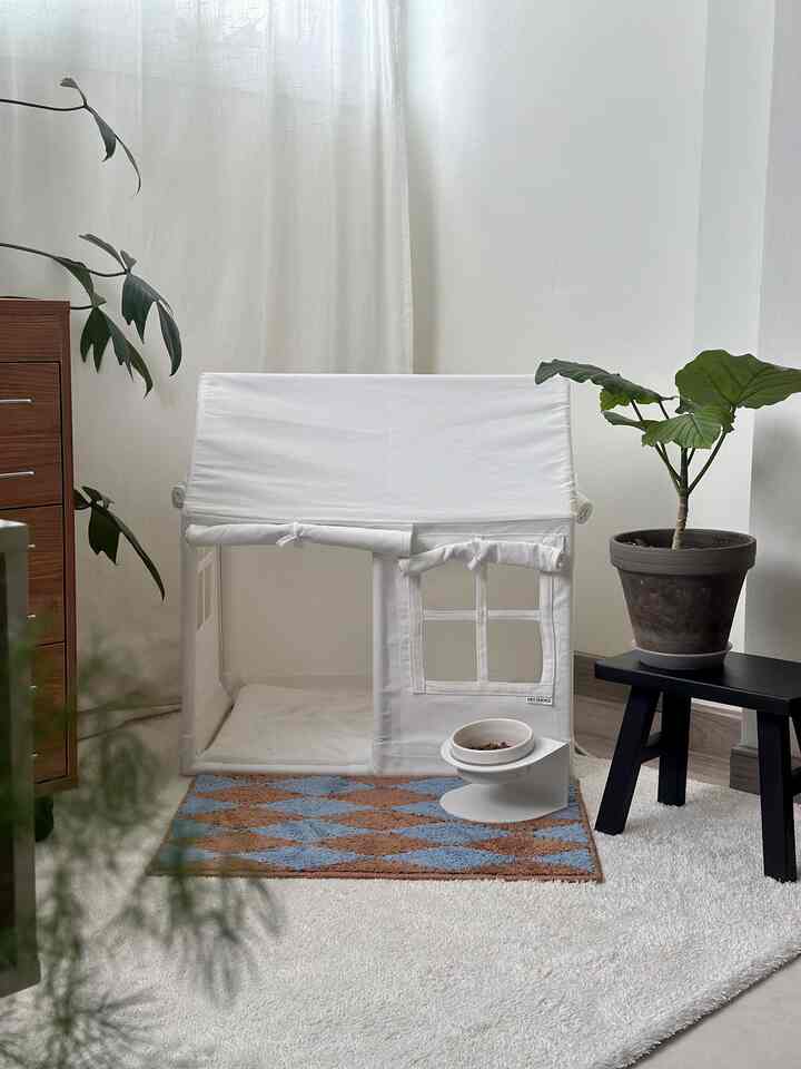 ホワイトトーンのリビングの一角に配置された犬用のホワイトペットハウス、ブラウンとブルーのチェック柄ラグ、緑豊かな植物が調和した雰囲気のあるインテリア