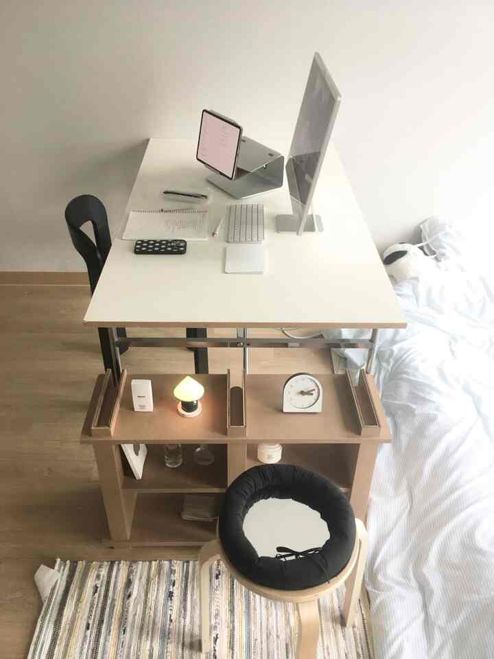 白と木目調が調和するワンルーム寝室、パソコンとタブレットが置かれたデスクとクッション付きスツールが配されたモダンな在宅勤務空間