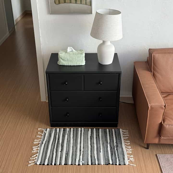 ナチュラルな木目の床とブラウンソファがあるモダンなリビング、黒のドレッサーに白いテーブルランプ、モノトーンのストライプラグが配置された印象的な空間