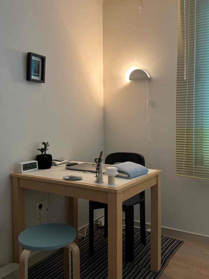 ナチュラルカラーの小さな部屋のホームオフィス、木製デスクと黒い椅子に壁の照明が配された落ち着いたインテリア