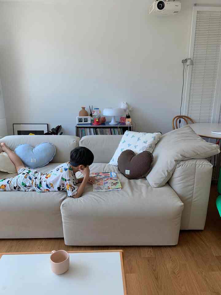 柔らかなベージュの色調が特徴のリビングルーム、ハート型クッションが置かれた大きなベージュのソファの上で子供が本を読んでいる、温かみのある空間