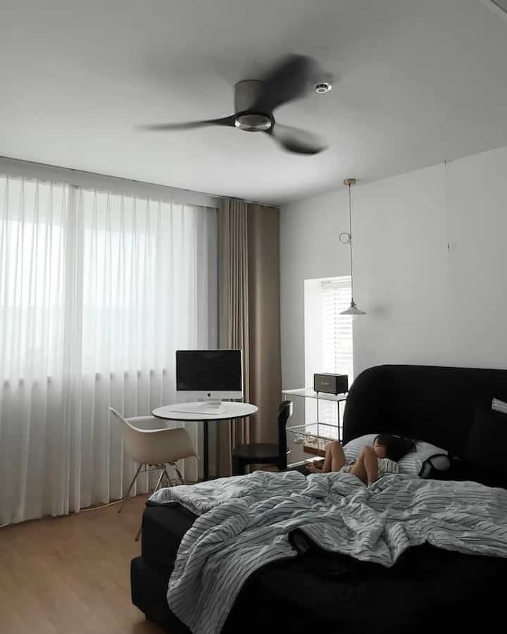 白と黒のナチュラルトーンの寝室、中央に回転するシーリングファン、窓際に丸テーブルと椅子が配置された落ち着いた雰囲気の空間