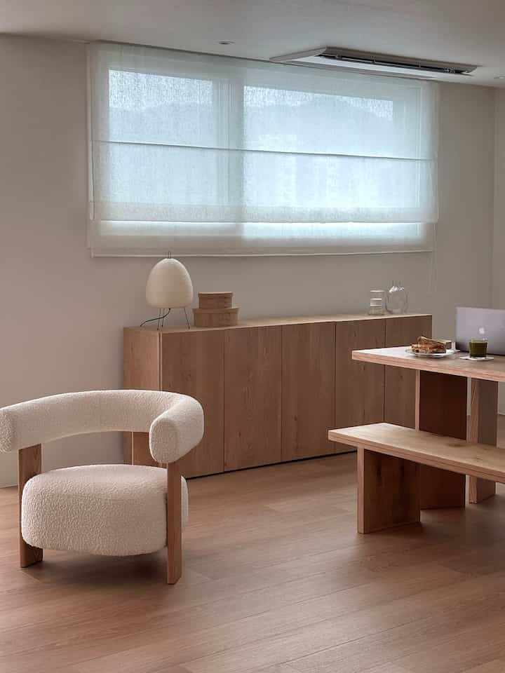 A simple natural-toned living room with white walls and brown wooden furniture, featuring a curved armchair and dining bench creating a cozy atmosphere