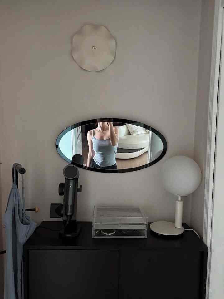 白い壁と黒いキャビネットがあるモダンな玄関、楕円形の鏡と特徴的な時計、テーブルランプが配置された雰囲気のあるインテリア