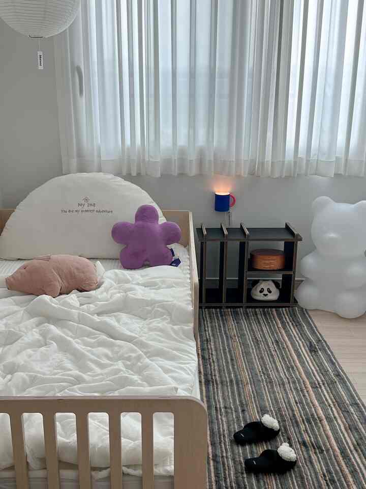 白い壁とカーテン、ナチュラルな木製ベッドにパープルのクッションが配された、モダンで自然な雰囲気の子供部屋