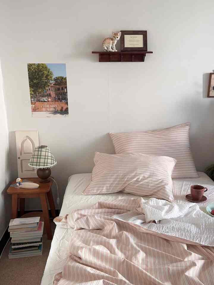 ナチュラルなベージュ色調の寝室、ピンクのストライプ寝具と小さな木製棚に猫のオブジェが配置されたシンプルな空間