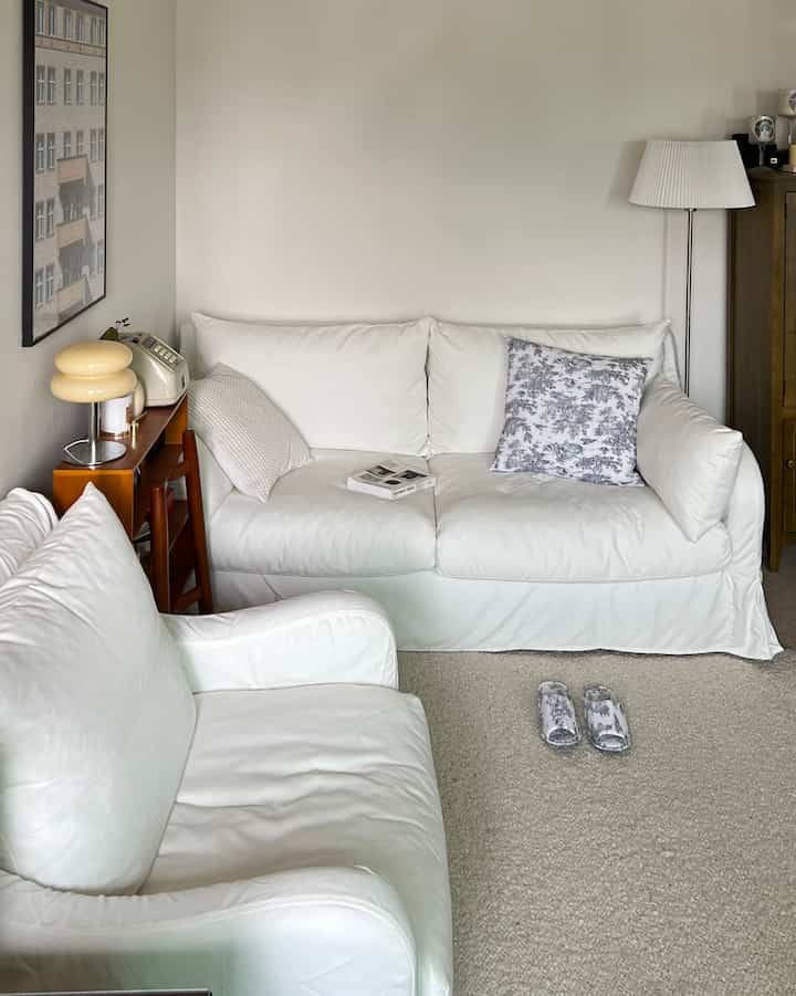 White-toned living room featuring a central white sofa and armchair, with a floor lamp and wooden side table on the right, creating a simple atmosphere