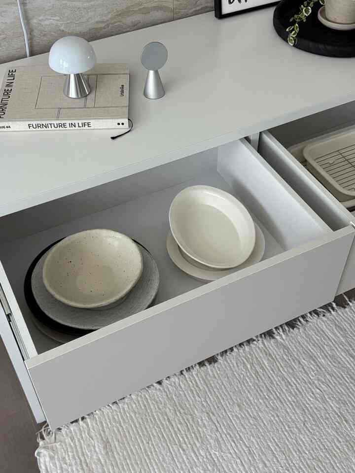 Minimal kitchen in white and beige tones featuring a neatly organized drawer with dishware and a white rug