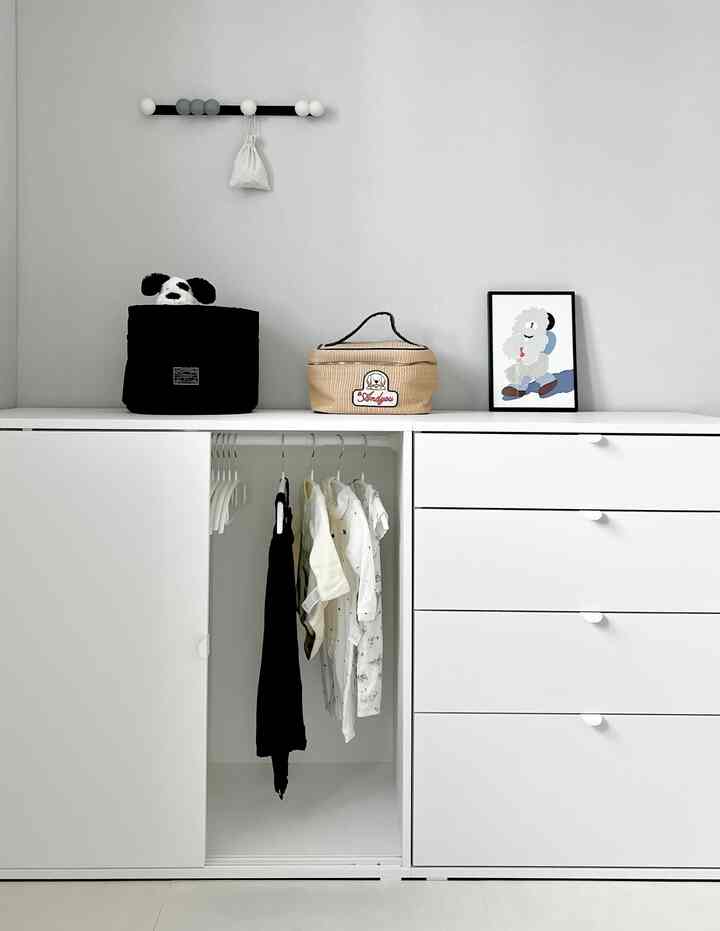 白を基調とした子供部屋、中央に洋服ダンスがあり、赤ちゃんの服と小物が整えられたモダンな空間