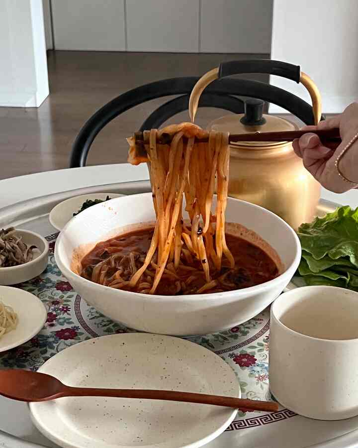 白色調のダイニングテーブルに置かれた皿と椀、木製のカトラリー、辛いスープの韓国料理風ヌードルが配置された雰囲気のある空間