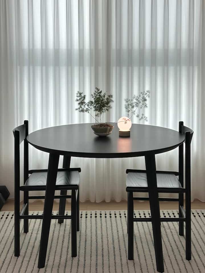 A simple modern living room featuring a black round dining table and two chairs against white curtains and a striped rug background