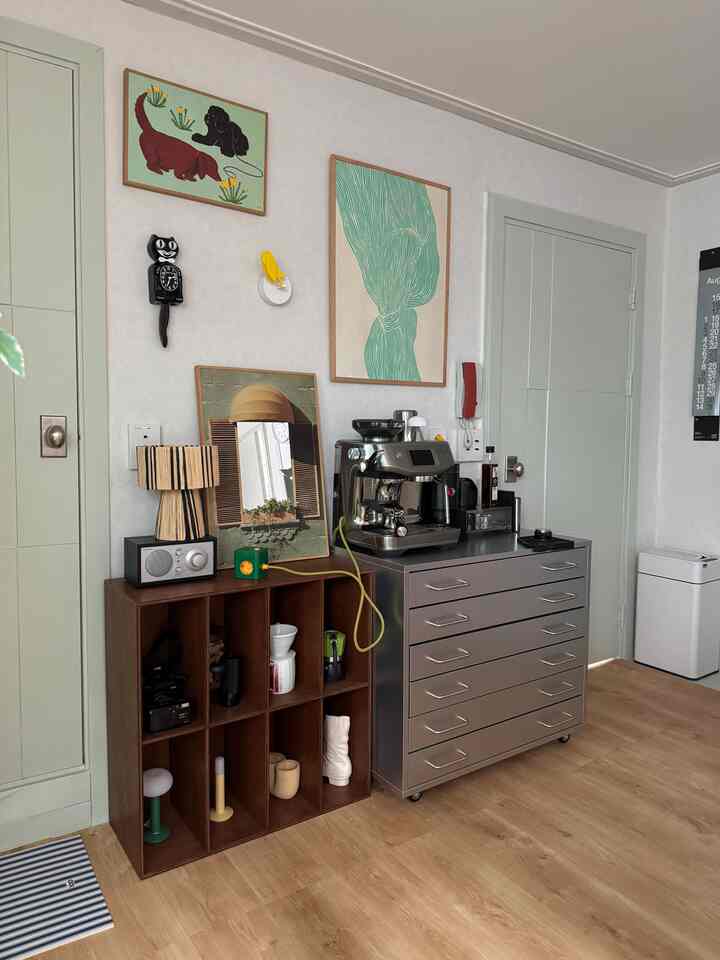 ブラウンの木目調の床とグリーンが調和したリビング、コーヒーマシンとヴィンテージの壁時計が配置された雰囲気のあるおうちカフェ