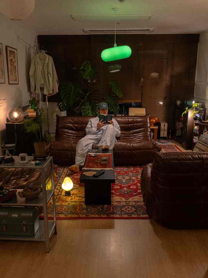 暖かいナチュラルトーンのリビング、ヴィンテージレザーソファと多彩なラグ、照明とブラインドが調和した雰囲気のあるインテリア