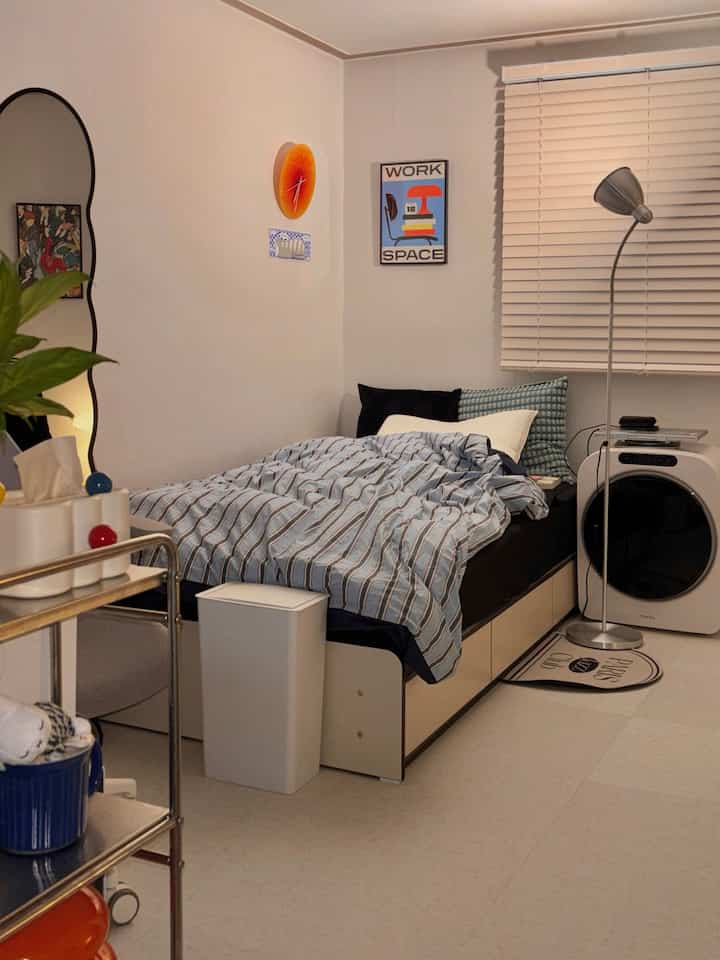 アイボリーとブラックのトーンが特徴のワンルーム寝室、ベッドフレームやブラインド、フロアランプが置かれた雰囲気のあるインテリア