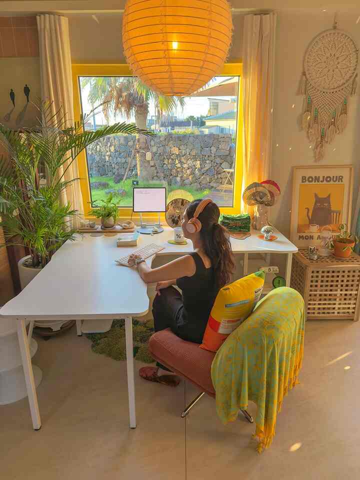 Warm natural-tone living room home office featuring large yellow-framed window, linen curtains, and plants creating a cozy atmosphere