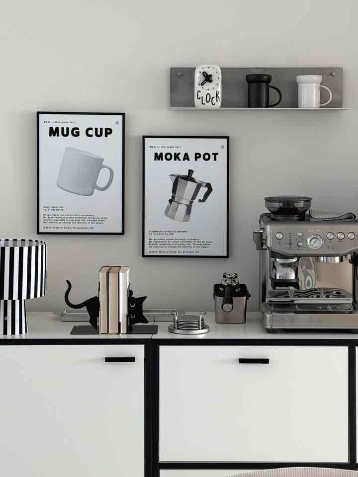 黒と白のモノトーンのキッチンスペース、ポスターとコーヒーマシン、壁棚、収納キャビネットが配置された雰囲気のあるおうちカフェスタイルのインテリア