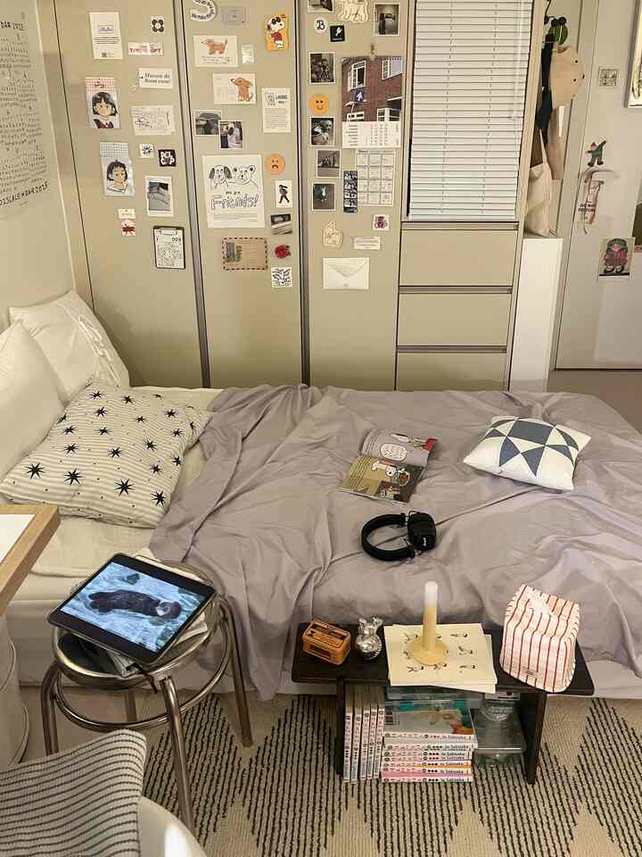 ベージュとグレーのモノトーンの一人暮らし寝室、ベッドとナイトテーブル、壁に様々なポスターとブラインドが配置された雰囲気のあるインテリア