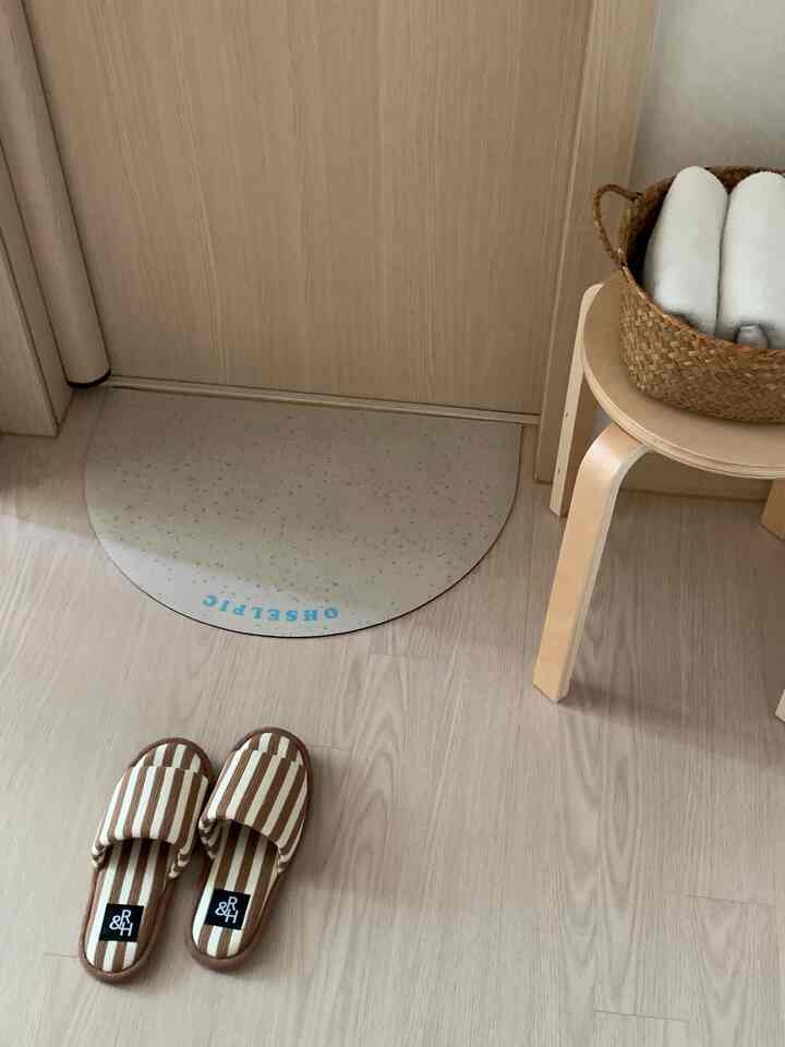 ナチュラルな木目床と木製スツールがある小さくてすっきりとした玄関、扉前に半円形の玄関マットとストライプのルームシューズが配置されている