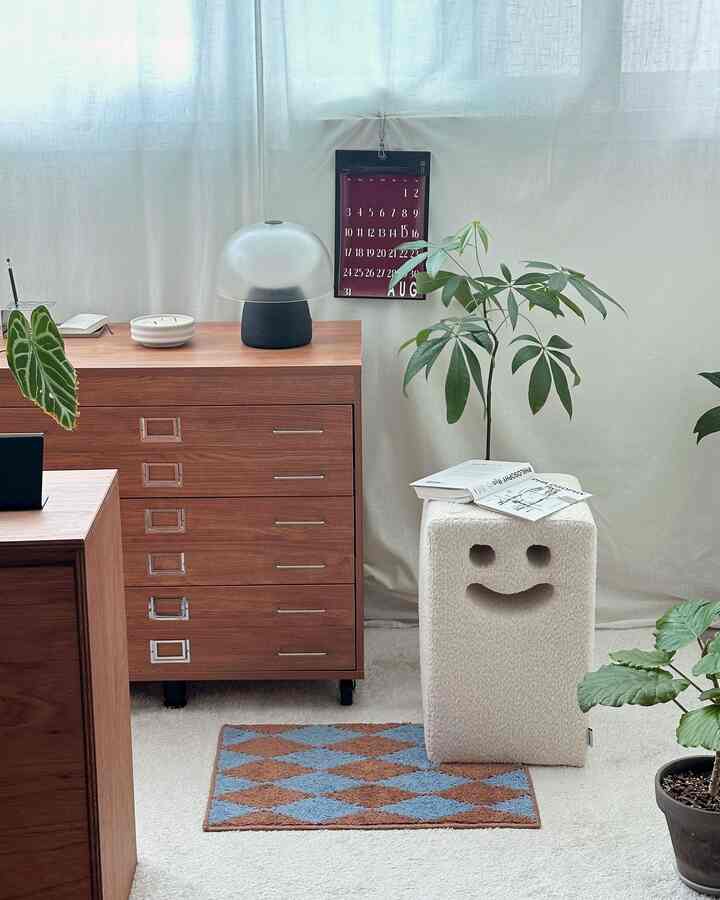 ホワイトのカーテンとカーペットが広がる子供部屋、木目調の収納チェストと笑顔のスツール、植物が配された明るく居心地の良い空間